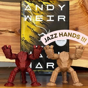 May include: Two 3D-printed figures, one maroon and one tan, stand in front of a book titled "The Martian" by Andy Weir. The figures have a textured appearance and are posed with their arms raised. The text "JAZZ HANDS !!!" is above the figures.