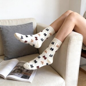 May include: Off-white socks patterned with cat faces in black, grey, brown, and peach. The socks are mid-calf length. The person wearing them is relaxing on a sofa. The socks are a fun and quirky accessory.