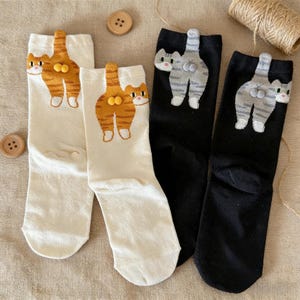 Op de afbeelding: Vier paar sokken met kattenontwerpen. Twee paar zijn gebroken wit met oranje tabby kattenafbeeldingen en twee zijn zwart met grijze en witte kattenafbeeldingen. De kattenontwerpen staan op de achterkant van de sokken.