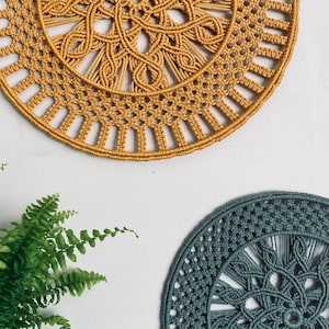 Op de afbeelding: Twee macramé wandkleden. De bovenste is mosterdgeel, met een ingewikkeld cirkelvormig ontwerp. De onderste is blauwgroen, met een vergelijkbaar patroon. Beide worden weergegeven tegen een witte muur, met een varen in de linker benedenhoek.