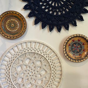 May include: A collection of handmade wall decor, including a navy blue macrame hanging, a large cream-colored macrame doily, and two decorative wooden plates with intricate painted designs. The items are displayed against a white wall.