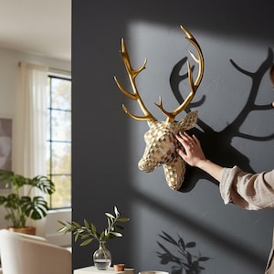 Könnte beinhalten: Goldfarbene Hirschkopf-Wanddekoration mit detailliertem Geweih. Der Hirschkopf hat ein mosaikartiges Muster in Gold- und Weißtönen. Die Wand ist dunkelgrau.
