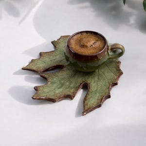 May include: A small, green ceramic cup filled with coffee sits on a leaf-shaped ceramic coaster. The coaster is green with brown accents, and the cup has a matching green handle. The coffee has a foamy top.