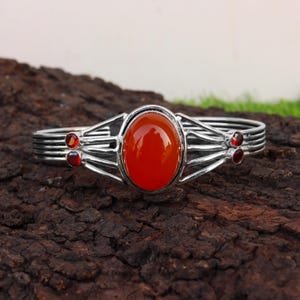 Op de afbeelding: Zilverkleurige armband met een grote, ovale, oranjerode edelsteen in het midden. De armband heeft een reeks parallelle banden en decoratieve accenten, met kleinere rode edelstenen aan weerszijden van de centrale steen. Op een donker, getextureerd oppervlak.