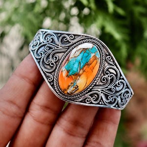 Könnte beinhalten: Ein silberner Armreif mit einem ovalen Stein in leuchtendem Orange und Türkisblau. Das Metallband ist mit komplizierten, erhabenen Schnörkelmustern verziert und weist eine gedrehte Kordelkante auf, die den zentralen Stein umschließt.