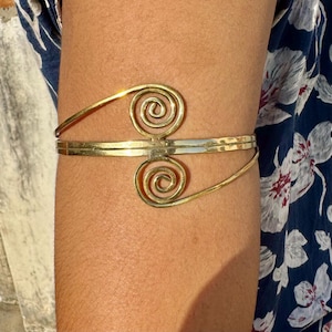 Könnte beinhalten: Ein goldfarbener Armreif mit Spiralmuster. Der Armreif hat zwei spiralförmige Akzente, die durch ein Band verbunden sind, das den Arm umschließt. Das Design ist schlicht und elegant, passend für verschiedene Anlässe.
