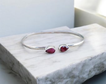 Puede incluir: Una pulsera de plata con dos gemas rojas en forma de lágrima. La pulsera se exhibe sobre una superficie de mármol blanco y gris, mostrando su elegante diseño y el color vibrante de las piedras.
