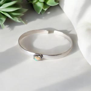 Könnte beinhalten: Ein silbernes Armreif-Armband mit einem ovalen Opal-Edelstein. Das offene Armband hat eine glatte, polierte Oberfläche. Der Opal ist in einer strukturierten Lünette gefasst. Der Edelstein zeigt ein Farbspiel, darunter Weiß, Gelb und Grün.