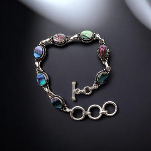 Könnte beinhalten: Silberarmband mit ovalen Abalone-Muschelsteinen in Blau-, Grün- und Lilatönen. Das Armband hat einen Knebelverschluss und ist vor einem dunklen Hintergrund platziert. Das Armband ist ein Schmuckstück.