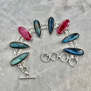 May include: A silver bracelet featuring a series of teardrop-shaped gemstones in shades of red, blue, and green. The gemstones are connected by silver chain links and a toggle clasp. The bracelet is displayed on a gray surface.