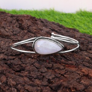 Könnte beinhalten: Ein silbernes Armreif-Armband mit einem tropfenförmigen, hellrosa Edelstein. Der Edelstein ist in einen silbernen Rahmen gefasst, und das Armband hat ein schlichtes, offenes Design. Das Armband wird auf einer strukturierten, braunen Oberfläche präsentiert.