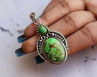 May include: A silver pendant with a large, oval-shaped green stone with brown veining, and a smaller, teardrop-shaped green stone above it. The pendant has a decorative silver border with small beaded accents.