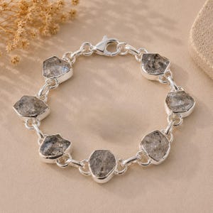 Peut inclure: Un bracelet à chaîne en argent composé de six pierres grises translucides et irrégulières, serties dans des chatons en argent poli. Le bracelet est fermé par un fermoir mousqueton en argent, reliant chaque pierre par des anneaux argentés.