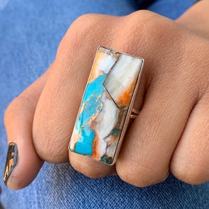 May include: A rectangular silver ring featuring a mosaic of turquoise, white, orange, and gold-colored stone. The ring's design is a striking contrast of colors and textures, showcasing a unique, eye-catching piece of jewelry.