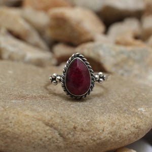 Könnte beinhalten: Ein Silberring mit einem tropfenförmigen, tiefroten Edelstein. Der Edelstein ist von einem gedrehten Seildesign umgeben und auf einem Silberband mit dekorativen Elementen gefasst. Der Ring wird auf einer neutral getönten Steinoberfläche präsentiert.