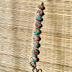 Può includere: Un bracciale in argento con pietre ovali nei toni dell'arancione, turchese e marrone. Le pietre sono incastonate in montature individuali in argento e collegate da maglie d'argento. Il bracciale è esposto su un tappetino intrecciato.