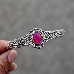 Puede incluir: Un brazalete de tono plateado que presenta una piedra cabujón ovalada de color rosa vibrante en el centro. La banda de metal está decorada con intrincados relieves de volutas y detalles tipo cuerda que rodean la gema rosa central.