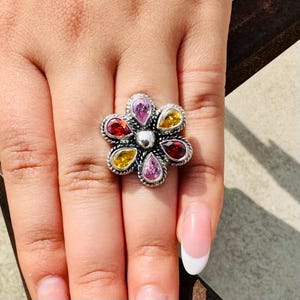 Könnte beinhalten: Ein silberner Blumenring mit farbigen Edelsteinen. Der Ring hat eine zentrale silberne Perle, die von sechs tropfenförmigen Steinen in Rot-, Gelb- und Rosatönen umgeben ist. Das Band des Rings hat ein strukturiertes, seilartiges Design.