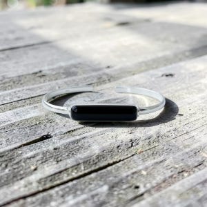 Könnte beinhalten: Ein silbernes Armreif-Armband mit einem rechteckigen schwarzen Stein als Mittelstück. Das offene Armband liegt auf einer verwitterten Holzoberfläche und zeigt sein minimalistisches Design und die kontrastierenden Farben. Die elegante, moderne Ästhetik des Armbands wird hervorgehoben.