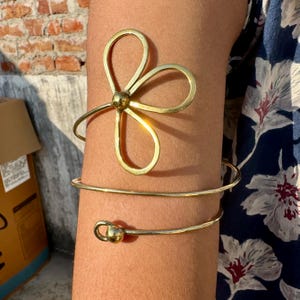 Könnte beinhalten: Goldfarbene Armspange mit floralem Design und Spiralarmband. Der obere Teil der Spange hat eine blütenartige Form mit drei gebogenen Blütenblättern. Das untere Band umschlingt den Arm. Schmuck.