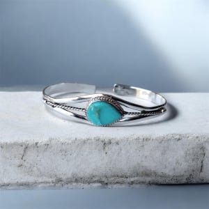 Könnte beinhalten: Ein silbernes Armreif-Armband mit einem tropfenförmigen Türkisstein. Das Armband hat ein geflochtenes Seil-Design auf beiden Seiten des Steins. Der Türkisstein hat eine leuchtende blaugrüne Farbe. Das Armband wird auf einer weißen Oberfläche präsentiert.