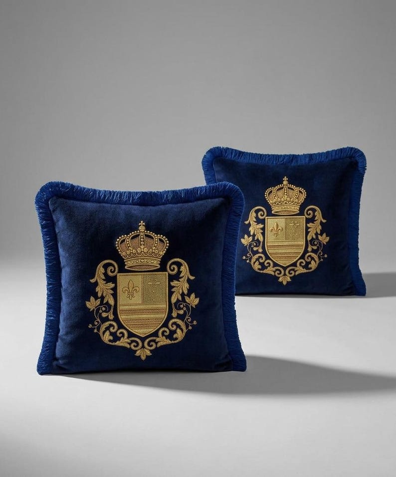 Puede incluir: Dos cojines cuadrados de terciopelo azul marino con escudos bordados en hilo dorado que presentan un motivo de corona y flor de lis. Cada coj&iacute;n est&aacute; rematado con un fleco azul marino a juego en los bordes, aportando un acabado texturizado.