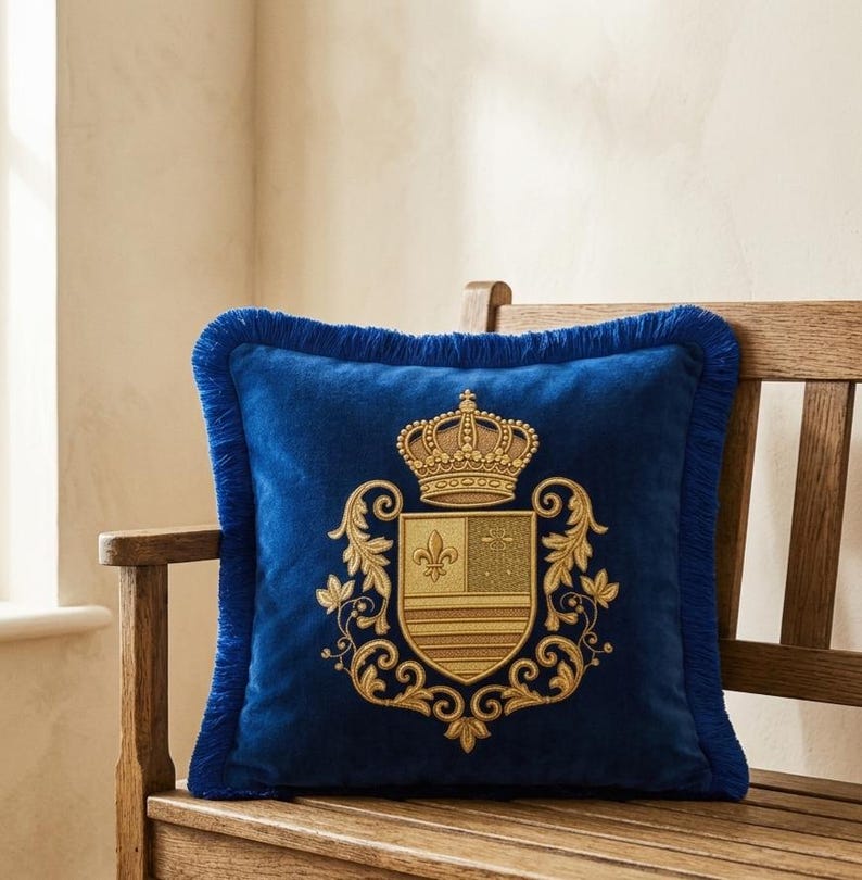 Puede incluir: Un coj&iacute;n cuadrado de terciopelo azul real con un escudo bordado en oro que presenta una corona y un motivo de flor de lis. El coj&iacute;n est&aacute; rematado con un borde de flecos azul a juego en los cuatro lados, creando un aspecto texturizado.