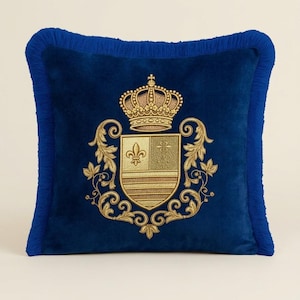 Puede incluir: Un coj&iacute;n cuadrado de terciopelo azul real con un escudo bordado en hilo dorado que presenta una corona y un motivo de flor de lis. El coj&iacute;n est&aacute; rematado con un fleco azul a juego en los cuatro bordes, creando un borde texturizado.