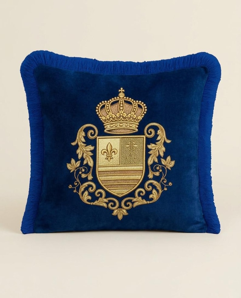 Puede incluir: Un coj&iacute;n cuadrado de terciopelo azul real con un escudo bordado en hilo dorado que presenta una corona y un motivo de flor de lis. El coj&iacute;n est&aacute; rematado con un fleco azul a juego en los cuatro bordes, creando un borde texturizado.
