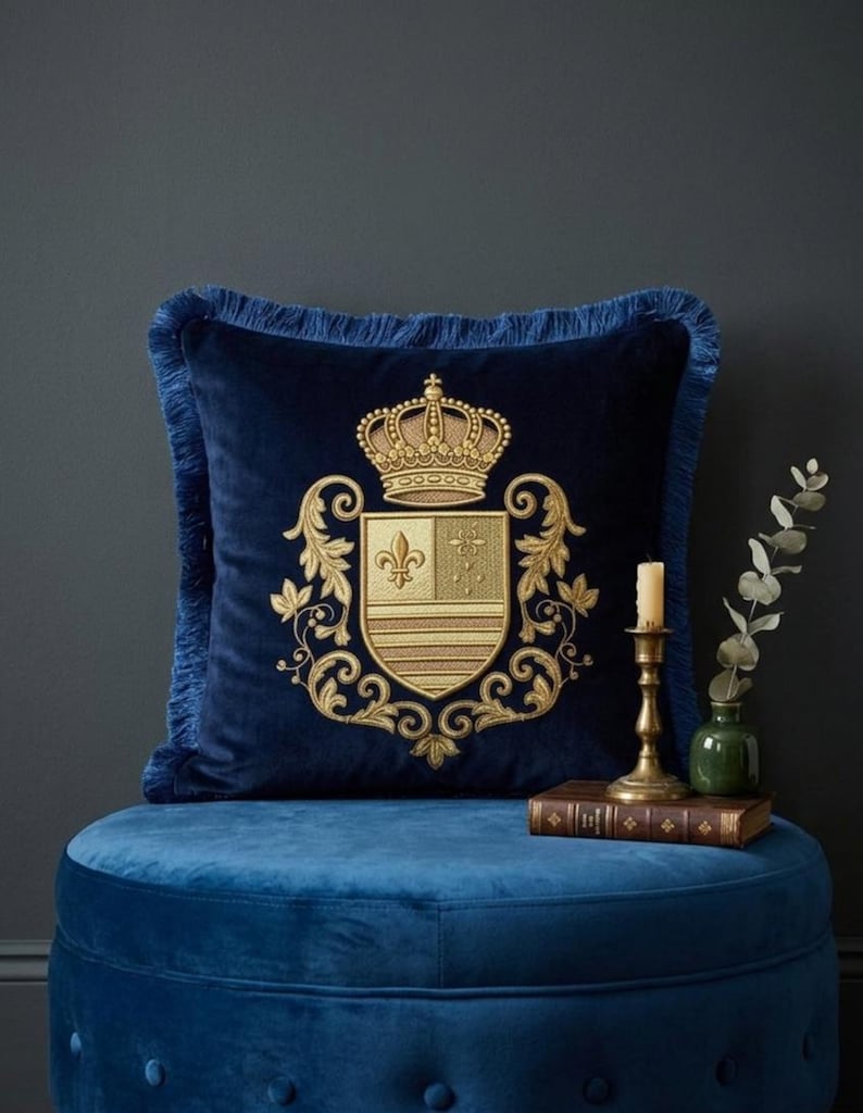 Puede incluir: Un coj&iacute;n cuadrado de terciopelo azul marino con un escudo bordado en hilo dorado que presenta una corona y un motivo de flor de lis. El coj&iacute;n est&aacute; rematado con un fleco a juego en color azul marino en los cuatro bordes, creando un acabado texturizado.