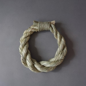 May include: A circular wreath crafted from light tan, dried natural straw fibers. The straw is tightly braided into a thick, continuous loop, with a distinct section at the top secured by a neat, horizontal wrapping of the same straw material.