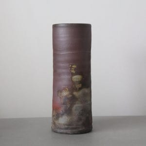 May include: A tall, cylindrical ceramic vase with a dark burgundy exterior. The vase has a textured surface with areas of gold, red, and gray. The vase is a decorative item for the home.