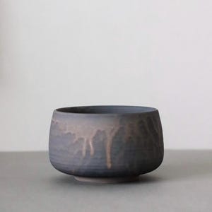 May include: A handcrafted, dark blue ceramic bowl with a textured exterior. The bowl features a unique design with a drip-like pattern in a lighter, earthy tone. The bowl has a rounded shape and sits on a small, circular base.