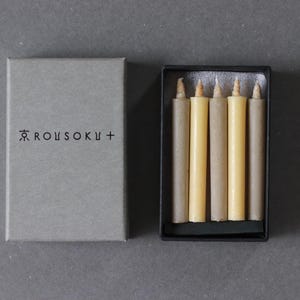 以下が含まれることがあります： 黒いトレイに入った、自然な質感のクリーム色のろうそく6本セット。ろうそくは「ROUSOKU +」の文字が印刷された灰色の箱に入っています。背景は無地のグレーです。