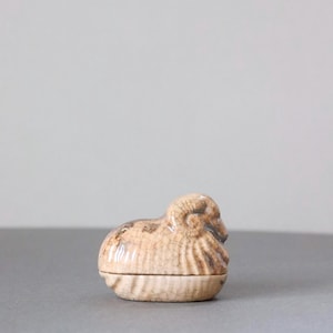 Op de afbeelding: Een klein, beige keramieken snuisterijendoosje in de vorm van een ram. Het oppervlak heeft een opvallende craquelé glazuurafwerking met donkerbruine accenten die de textuur van de gekrulde hoorns en de wollige vacht benadrukken, bestaande uit een basis en deksel.