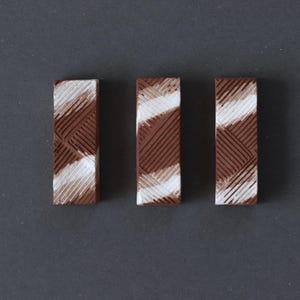 Könnte beinhalten: Drei rechteckige, strukturierte Objekte mit einem braunen und weißen diagonalen Muster. Die Objekte sind vertikal vor einem dunkelgrauen Hintergrund angeordnet. Das Design zeigt diagonale Linien und einen helleren, weißen Bereich.