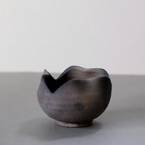 以下が含まれることがあります： 暗く土のような色の、手作りの小さなセラミックボウル。ボウルは、波状の縁と小さく丸みを帯びたベースを持つ、ユニークで有機的な形状をしています。外側は質感があり、自然で素朴な美しさを感じさせます。ボウルは装飾目的で使用される可能性があります。