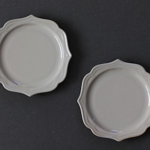 Può includere: Due piatti bianco sporco con bordo smerlato sono esposti su una superficie grigio scuro. I piatti hanno un design semplice ed elegante, con un bordo leggermente rialzato e una finitura liscia e opaca. I piatti sono rotondi con un bordo decorativo.