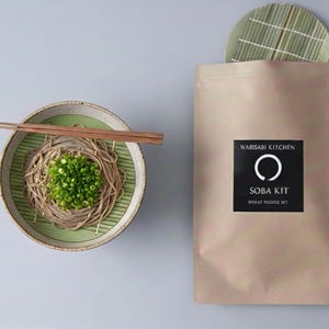 Puede incluir: Un kit de soba preparado con fideos de trigo, cebolletas y palillos, presentado en un cuenco de cerámica. Un paquete etiquetado como "Wabisabi Kitchen Soba Kit" está al lado del cuenco, con una esterilla de bambú encima.