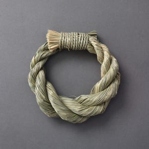 May include: A circular, woven wreath made of natural, light green and tan fibers. The wreath is tightly braided, with a thicker, wrapped section at the top. The background is a solid, neutral gray.