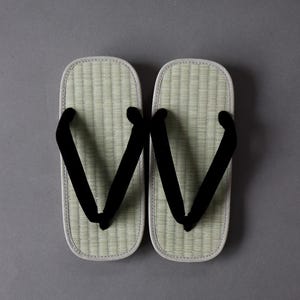 May include: A pair of traditional Japanese geta sandals. The sandals feature a light green woven straw footbed, a white border, and black fabric straps. The geta are set against a gray background.