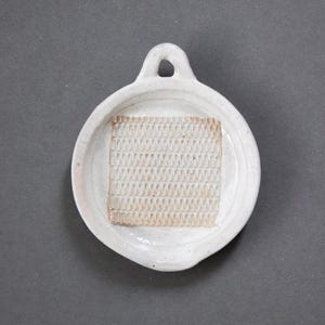 May include: A white ceramic grater with a handle and a textured surface. The grater has a round shape with a square, textured grating surface in the center. The handle has a hole for hanging.