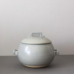 May include: A round, light gray ceramic jar with a lid and two side handles. The lid has a small handle on top. The jar sits on a gray surface with a white background. The jar is a handmade pottery piece.