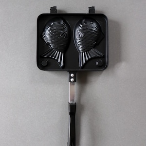 May include: A black metal taiyaki pan featuring two fish-shaped molds. The cooking surface is rectangular with a long handle attached by two silver rivets. The design includes detailed scales and fins on the fish molds, with a smooth black finish.