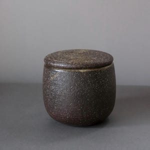 Pode incluir: Um pequeno pote de cerâmica castanho escuro com tampa a condizer. O pote tem uma superfície texturizada e uma forma arredondada. A tampa assenta perfeitamente, criando uma estética rústica. O pote é provavelmente usado para armazenamento.