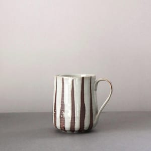 May include: A ceramic mug featuring a cylindrical body with a white base glaze and vertical dark brown painted stripes. The mug has a curved handle on the right side and a smooth, slightly tapered silhouette with a flat base and a rounded rim.