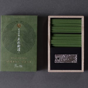 Puede incluir: Una caja de madera abierta y una caja verde de varillas de incienso. La caja verde de la izquierda tiene texto en relieve y un diseño circular. La caja abierta de la derecha contiene muchas varillas de incienso verdes y una placa de metal plateado.