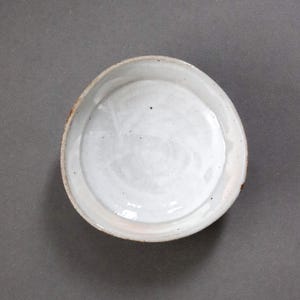 May include: A handmade, off-white ceramic dish with a slightly irregular shape. The interior is smooth and glossy, while the rim has a rustic, textured appearance. The dish is suitable for serving food or as a decorative piece.