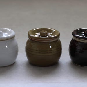 以下が含まれることがあります： 蓋付きの小さなセラミックジャーが3つ。ジャーは白、タン、濃い茶色です。各ジャーは丸みを帯びたボディと蓋の小さなノブが付いています。ジャーは、スパイスやその他の小さなアイテムを保管するためのものです。