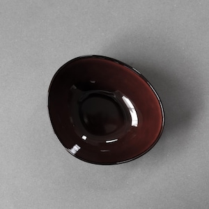 May include: A deep burgundy-colored, oval-shaped bowl. The bowl has a smooth, glossy finish and a dark interior. The bowl is set against a gray background. The bowl is likely made of glass or a similar material.